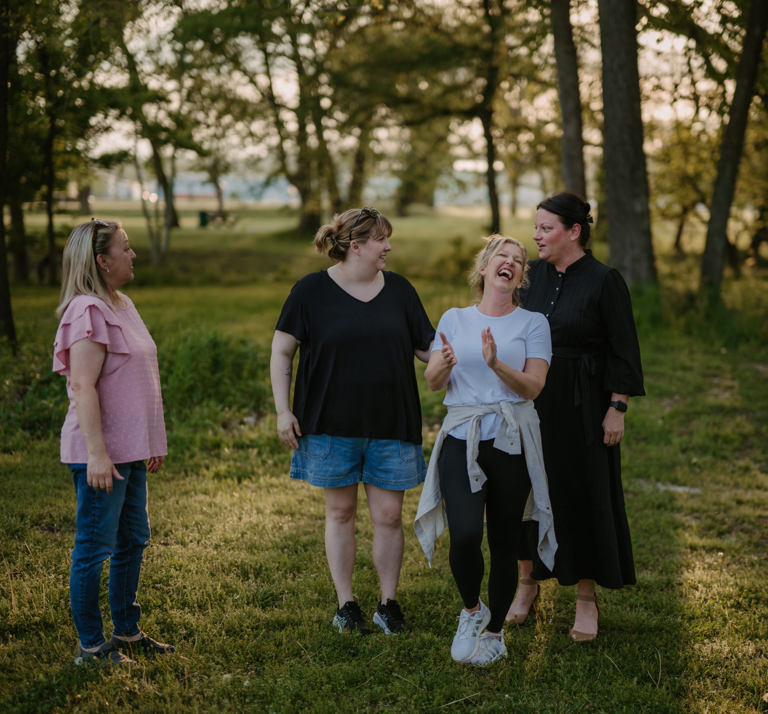 Four women stand together in a grassy park surrounded by trees, laughing and smiling in a candid moment. One woman in a white shirt throws her head back in laughter while the others look on with joy and warmth.