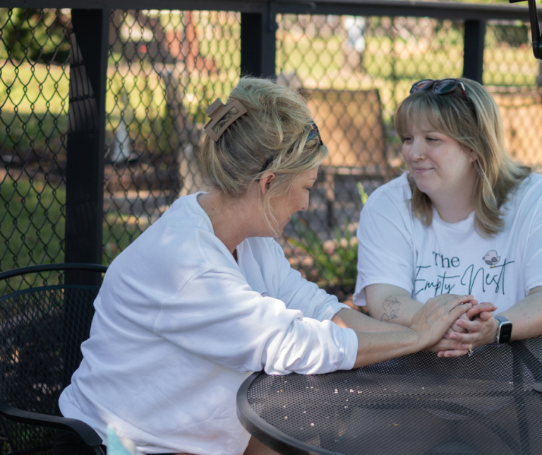 Mom & Daughter sitting at an outdoor table holding hands, smiling and talking — symbolizing honest connection between mother and daughter.