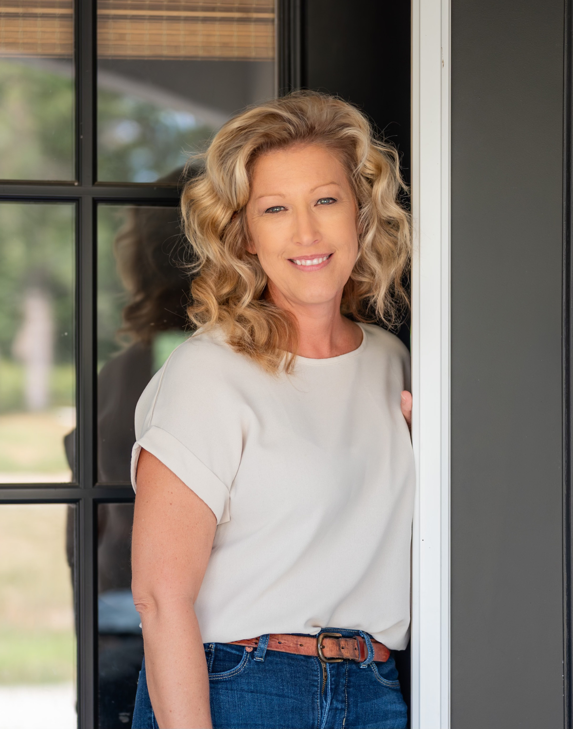 Mandy, a smiling blonde woman with curly hair, stands in a doorway wearing a cream top and jeans, looking warm and welcoming.