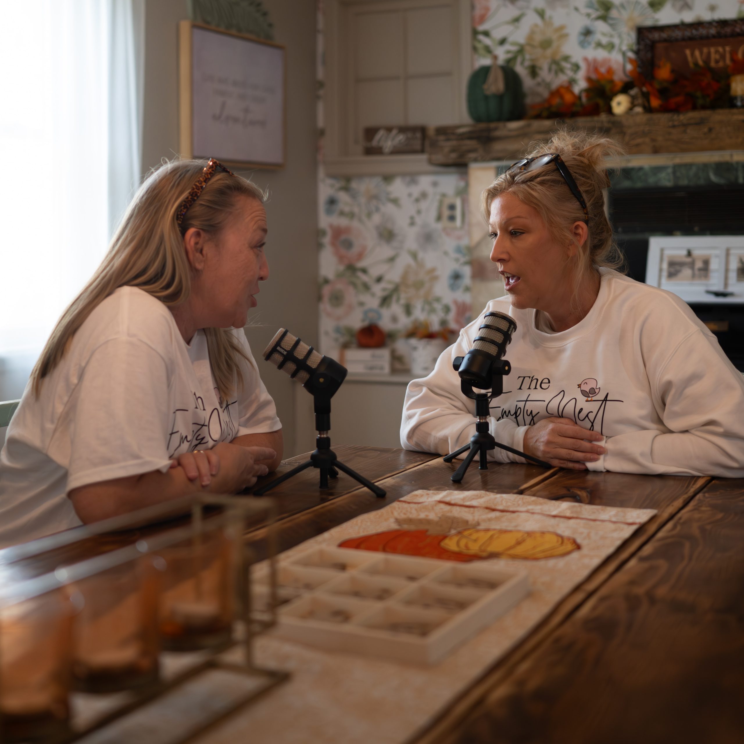 Mother and adult daughter recording The Empty Nest Mom podcast at a kitchen table with microphones, having an emotional conversation.