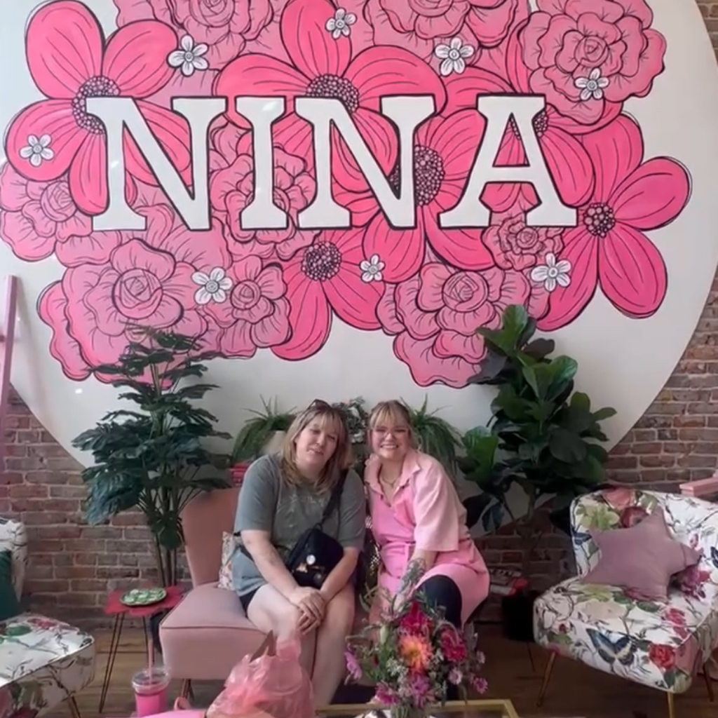Lauren posing with bakery owner Nina under pink floral wall sign near Kansas City