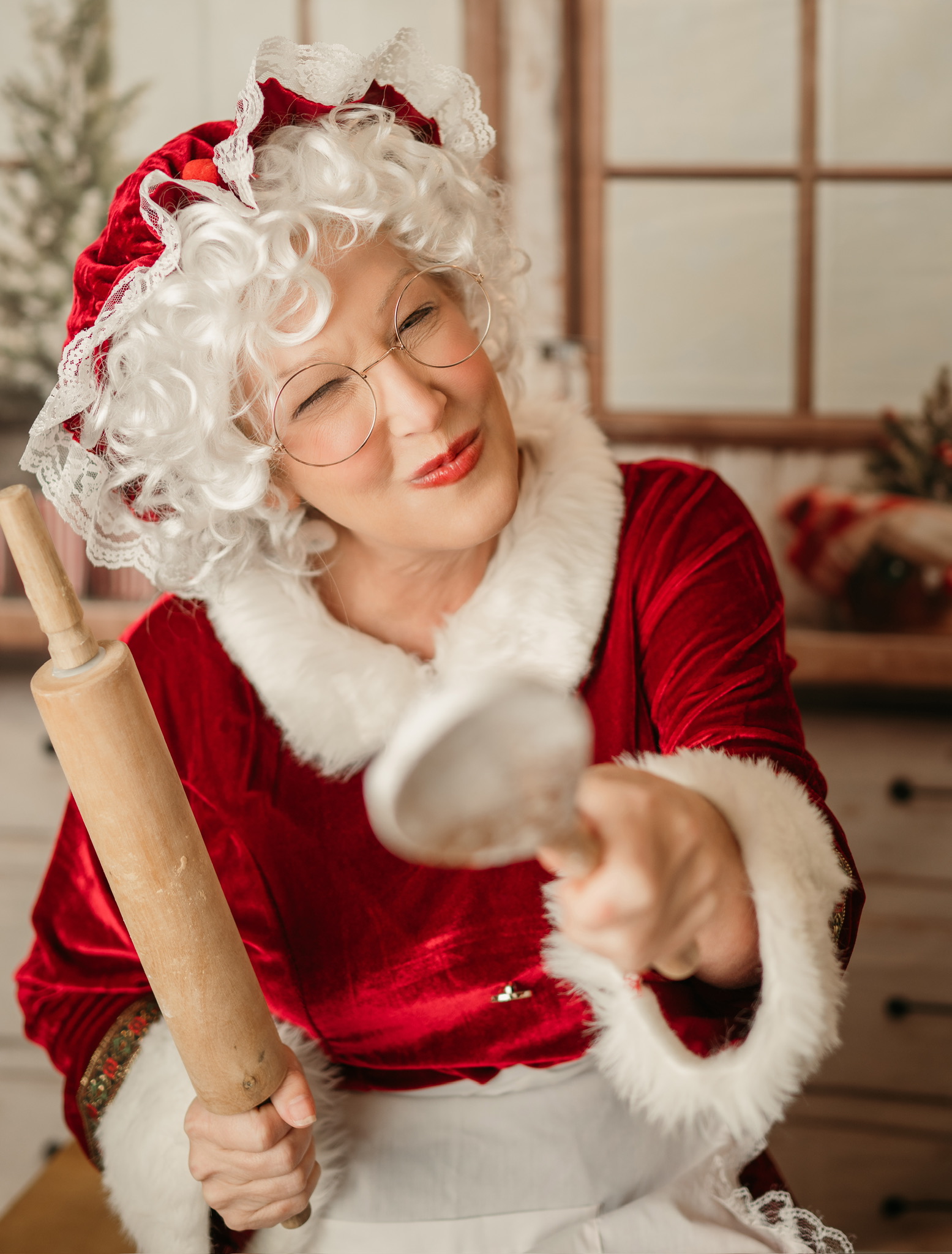 Mrs. Claus in a red velvet dress, white curly wig, and round gold glasses, pointing a wooden spoon and holding a rolling pin in her North Pole kitchen.