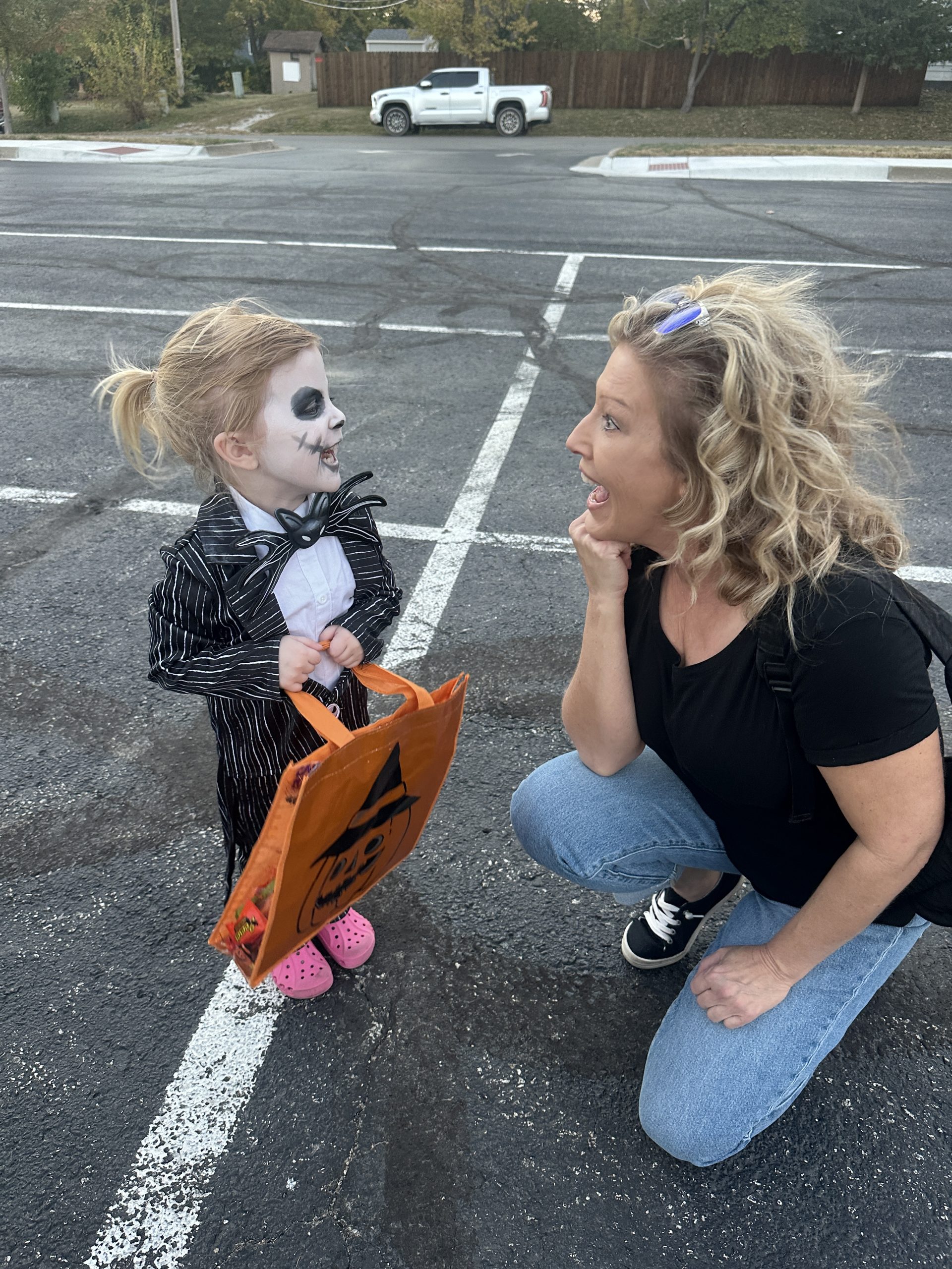 Grandmother kneeling in a parking lot with her little granddaughter in a Halloween costume and face paint, on a fall day